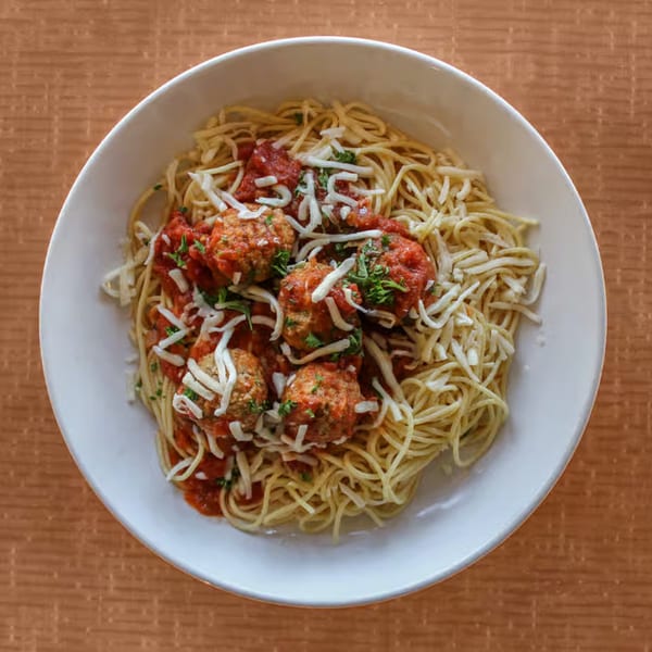 A white bowl of spaghetti and meatballs with red sauce, cheese, and parsley on a brown table.