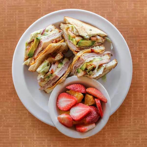 Four club sandwiches with avocado, cheese, and meat, served with a side of fresh strawberries.